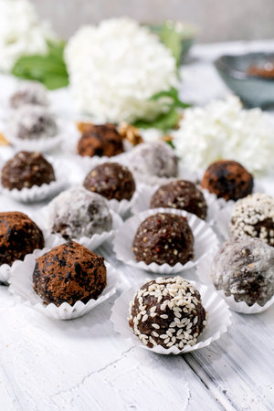 Various chocolate raw vegan balls truffles, coated with cocoa powder sesame seeds powdered sugar placed in paper cups on white wooden table with hydrangea flowers backgroundの写真素材