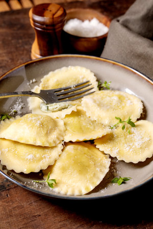 Fresh homemade ravioli pasta filled with cheese served on ceramic plate with fork garnished with herbs and parmesan cheese on rustic wooden tableの写真素材