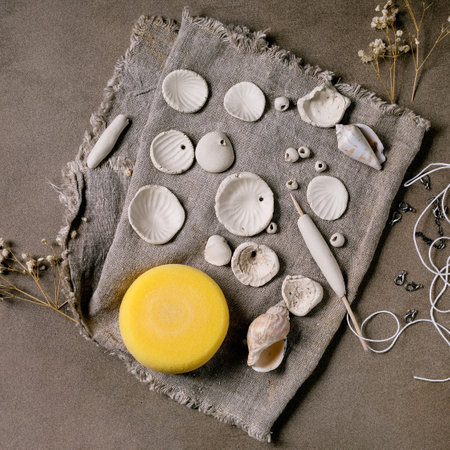 Ceramic workshop idea: handcrafted beads from clay or porcelain. Flat beads of raw white porcelain, tools on rough fabric. Small white flowers, jewelry findings, sea shells. Dark background, top view.の写真素材