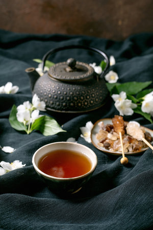 Traditional tea ceremony setup with cast iron teapot ceramic cup with hot tea sugar crystals jasmine flowers on dark linen backgroundの写真素材