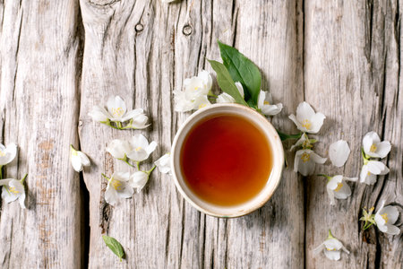 Cup tea surrounded by white jasmine flowers on rustic wooden background top view. Natural herbal drink with spring blossoms arrangementの写真素材