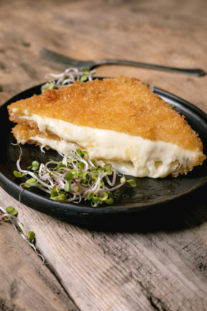Golden breaded cheese cutlet with melted filling served on black plate with microgreens garnish on rustic wooden table backgroundの写真素材