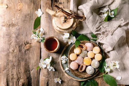 Vintage teapot and cup with colorful mochi sweets on rustic wooden table decorated with jasmine flowers and linen clothの写真素材