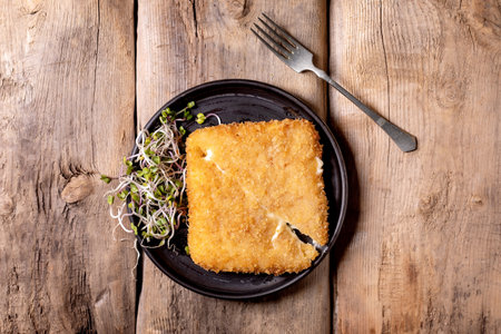 Golden breaded cheese cutlet served on black plate with microgreens fork beside on rustic wooden table top viewの写真素材