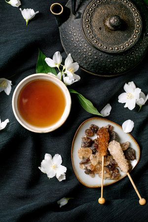 Traditional tea ceremony setup with cast iron teapot herbal tea cup rock sugar crystals wooden spoons and white jasmine flowers on dark linen backgroundの写真素材