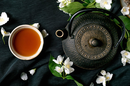Traditional cast iron teapot with cup tea surrounded by white jasmine flowers on dark green linen background. Top view flat lay composition for relaxation wellnessの写真素材