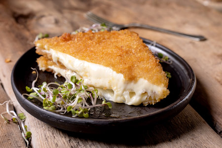 Golden crispy fried cheese with melted interior served on dark plate with microgreens wooden background comfort food conceptの写真素材