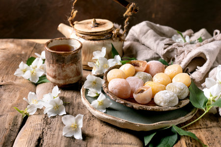 Ceramic plate with colorful mochi sweets tea cup teapot and white jasmine flowers on rustic wooden table with linen clothの写真素材