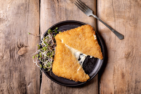 Golden fried cheese wedge melting on black pan with fresh microgreens fork on rustic wooden table backgroundの写真素材