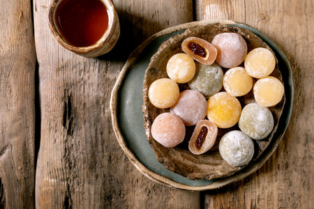 Colorful mochi balls with sweet filling served on ceramic plate alongside tea cup on rustic wooden table. Traditional Japanese confectionery dessert.の写真素材