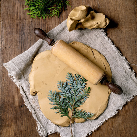 Decorating ceramics. A rolled slab of raw yellow clay with imprint of thuja branch lies on coarse fabric on wooden table. Eco decor. Rolling pin, tools, branches. Ceramic workshops. Top viewの写真素材