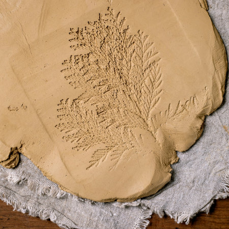 Decorating ceramics. Close up of rolled slab of raw yellow clay with imprint of thuja branch lies on coarse fabric on wooden table. Eco decor. Ceramic workshops, hand sculpting. Top viewの写真素材