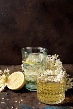 Two glasses elderflower beverages with fresh lemon half and white elderflower blossoms on rustic wooden background. Natural herbal refreshing drinks setup.の写真素材