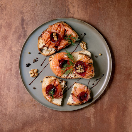 Sandwiches with thinly sliced salted salmon fillet, beetroot slices, walnuts, balsamic vinegar and fresh wheat bread on gray plate, fine dining. Brown background, top view. Colors of the year 2025.の写真素材