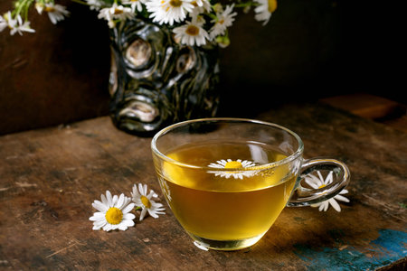 Glass cup herbal chamomile tea decorated with fresh daisy flowers on rustic wooden table. Natural wellness drink concept with floral arrangement background.の写真素材
