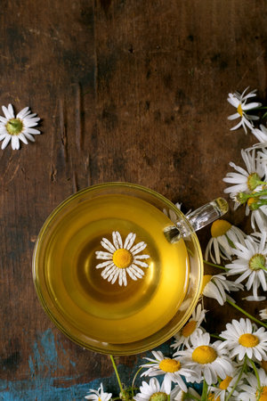 Glass cup chamomile tea with floating daisy flower surrounded by fresh chamomile flowers on rustic wooden background. Herbal tea concept natural remedy wellness.の写真素材