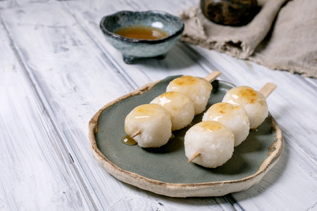 Sweet Japanese dango rice dumplings on wooden skewers served on ceramic plate with honey sauce traditional Asian dessert on white wooden tableの写真素材
