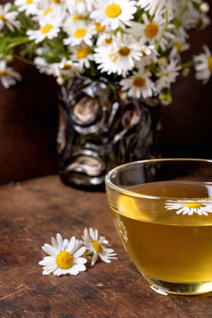 Glass cup herbal chamomile tea wooden table fresh white daisy flowers vase background. Natural wellness drink relaxation concept.の写真素材