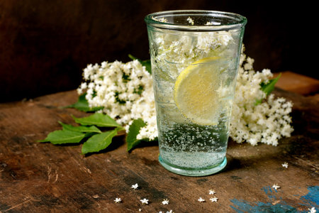 Glass sparkling water with lemon slice elderflower blossoms on rustic wooden table. Refreshing summer drink with natural floral decoration.の写真素材