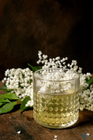 Golden elderflower cocktail in crystal glass with fresh white elderflower blossoms on rustic wooden background. Natural herbal drink garnished with flowers.の写真素材