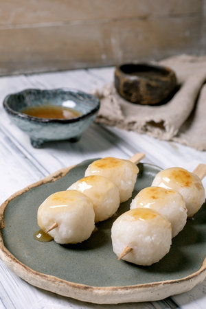 Traditional Japanese dango rice dumplings on wooden skewers served on ceramic plate with sweet mitarashi sauce and tea in rustic kitchen settingの写真素材