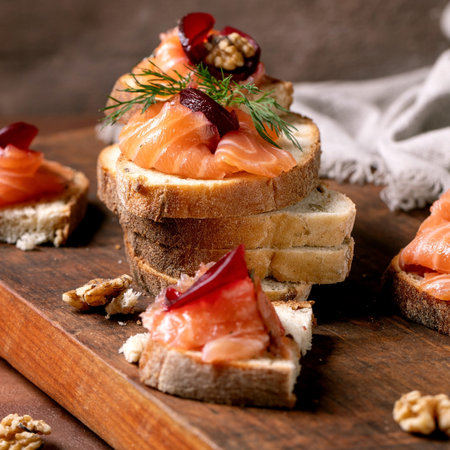 Sandwiches with thinly sliced salted salmon fillet, beetroot slices, walnuts, balsamic vinegar and fresh wheat bread in stack on wooden board. Brown background. Colors of the year 2025.の写真素材