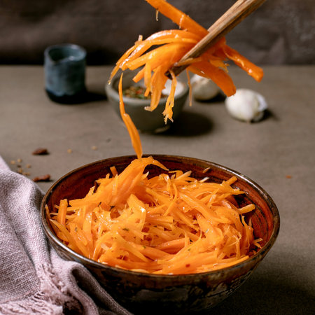 Korean and Asian cuisine. Man's hand uses chopsticks to pick up vegetable salad of marinated carrots with garlic from ceramic bowl. Seasoning in small bowls on Mocha Mousse tones table. Healthy eatingの写真素材