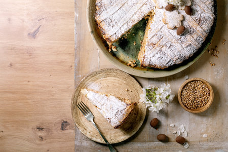 Traditional homemade pie Pastiera Napolitano dusted with powdered sugar. Slice served on ceramic plate. Decorated with wheat grains flowers and seeds on wooden table background.の写真素材