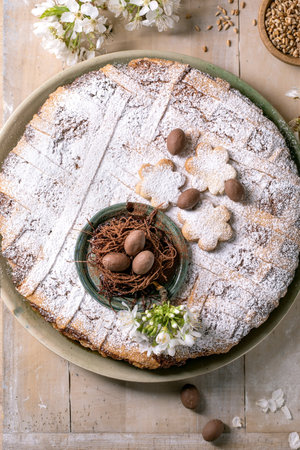 Homemade Easter cake Pastiera Napolitano with powdered sugar flower-shaped cookies chocolate eggs nest decoration. White spring flowers wooden table background rustic style.の写真素材