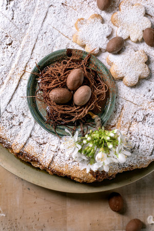 Traditional Easter dessert with chocolate eggs in nest on powdered traditional pie Pastiera Napolitano and white spring flowers on rustic background. Festive sweet composition.の写真素材