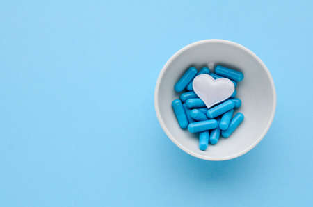 Blue medical capsules in white bowl and a white heart on a blue background. Concept of prevention and treatment of cardiovascular diseases, Valentine's day. Medical flat lay with copy space.の写真素材