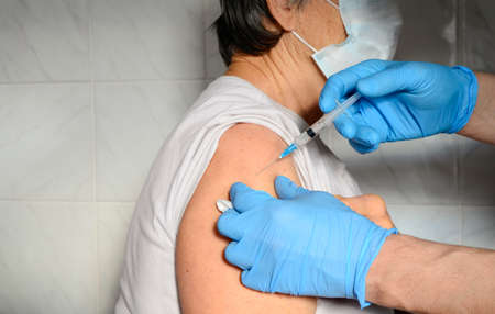 Elderly woman in medical mask receives vaccine in hospital. Blue-gloved hands inject vaccine into woman's forearm.の写真素材