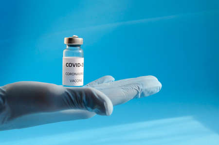 Hand in medical blue glove holding bottle of Covid 19 coronavirus vaccine on blue background with copy space and shadows. Vaccination concept, inscription Covid 19 coronavirus Vaccine.の写真素材
