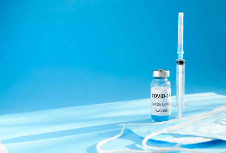 Glass bottle with inscription vaccine , medical masks and syringe on blue background with copy space and shadows. Concept of vaccination.の写真素材