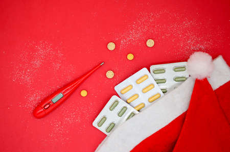 Top view of yellow and green medical capsules in a blister, medical pills and a red thermometer in a Santa hat on a red background. Medical Christmas concept.の写真素材