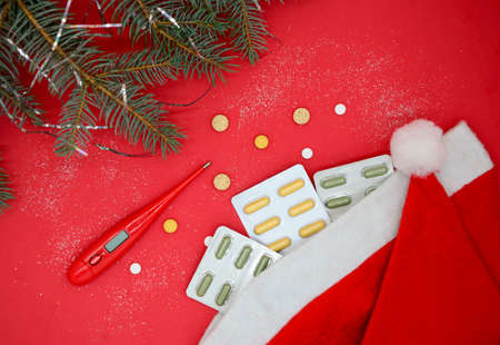 Top view of yellow and green medical capsules in a blister, medical pills and a red thermometer in a Santa hat on a red background. Medical Christmas concept.の写真素材