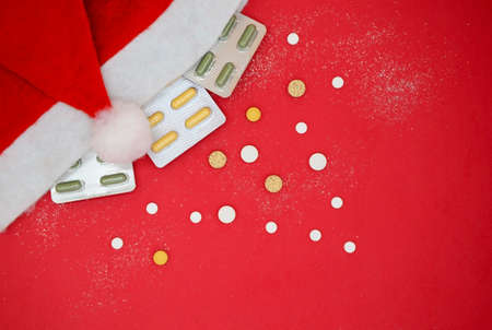 Top view of yellow and green medical capsules in a blister, medical pills in a Santa hat on a red background. Medical Christmas concept.の写真素材