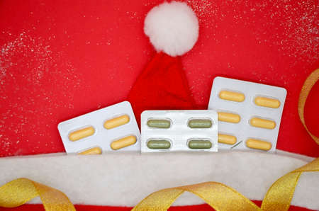 Top view of Christmas tree and yellow and green medical capsules in a blister, medical pills in a Santa hat and golden ribbon on red background. Medical Christmas concept, flat lay.の写真素材