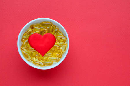 Yellow omega 3 capsules in white bowl and red heart on red background. Concept of proper nutrition, prevention of cardiovascular diseases, Valentine's Day. Medical layout with copy space.の写真素材
