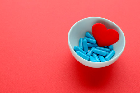 Blue medical capsules in white bowl and a red heart on red background. Concept of prevention and treatment of cardiovascular diseases, Valentine's day. Medical flat lay with copy space.の写真素材