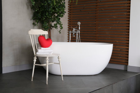 Modern bathroom interior design with concrete walls, wooden slats and white bathtub.vintage chair with towels and heart. Valentine's day concept, relaxation, massage, gifts, care, rest.の写真素材