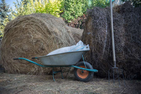 A garden cart with a bag of grain and a pitchfork near round bales of hay on the farm.の写真素材