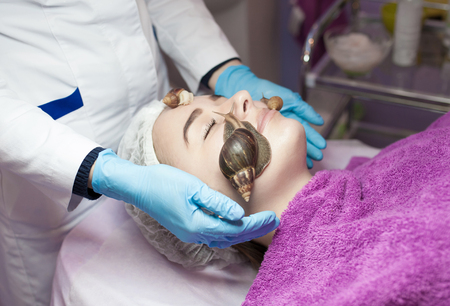 Young woman receiving snail facial massage. Snail on face. Cleaning procedure in spa salon. Blurred background.の写真素材