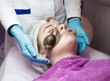 Young woman receiving snail facial massage. Snail on face. Cleaning procedure in spa salon. Blurred background.の写真素材
