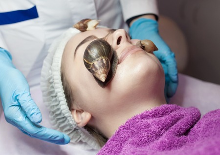 Young woman receiving snail facial massage. Snail on face. Cleaning procedure in spa salon. Blurred background.の写真素材
