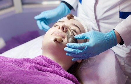 Young woman receiving snail facial massage. Snail on face. Cleaning procedure in spa salon. Blurred background.の写真素材