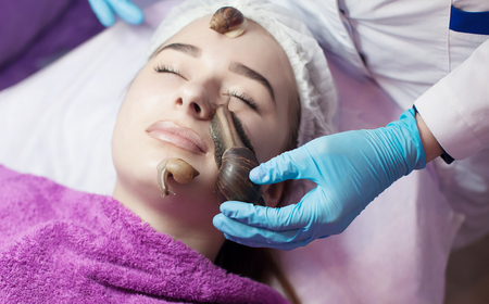 Young woman receiving snail facial massage. Snail on face. Cleaning procedure in spa salon. Blurred background.の写真素材