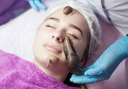 Young woman receiving snail facial massage. Snail on face. Cleaning procedure in spa salon. Blurred background.の写真素材