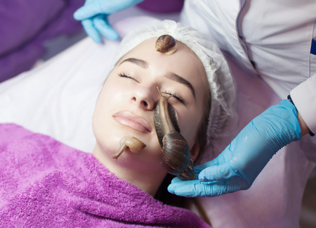 Young woman receiving snail facial massage. Snail on face. Cleaning procedure in spa salon. Blurred background.の写真素材
