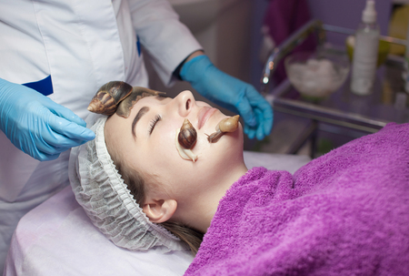 Young woman receiving snail facial massage. Snail on face. Cleaning procedure in spa salon. Blurred background.の写真素材